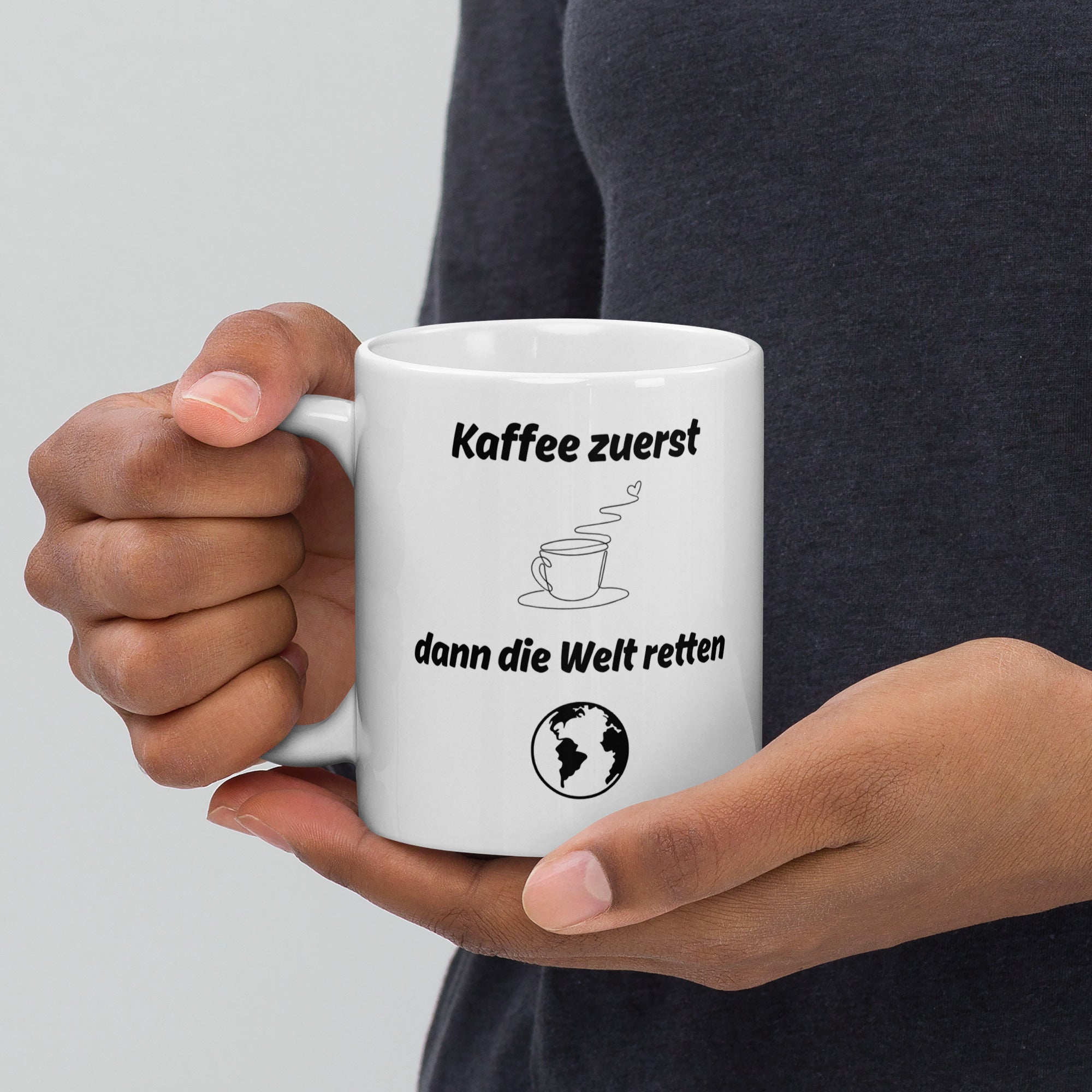 Eine Person hält eine weiße Kaffeetasse mit dem Spruch ‚Kaffee zuerst – dann die Welt retten‘ in der Hand.