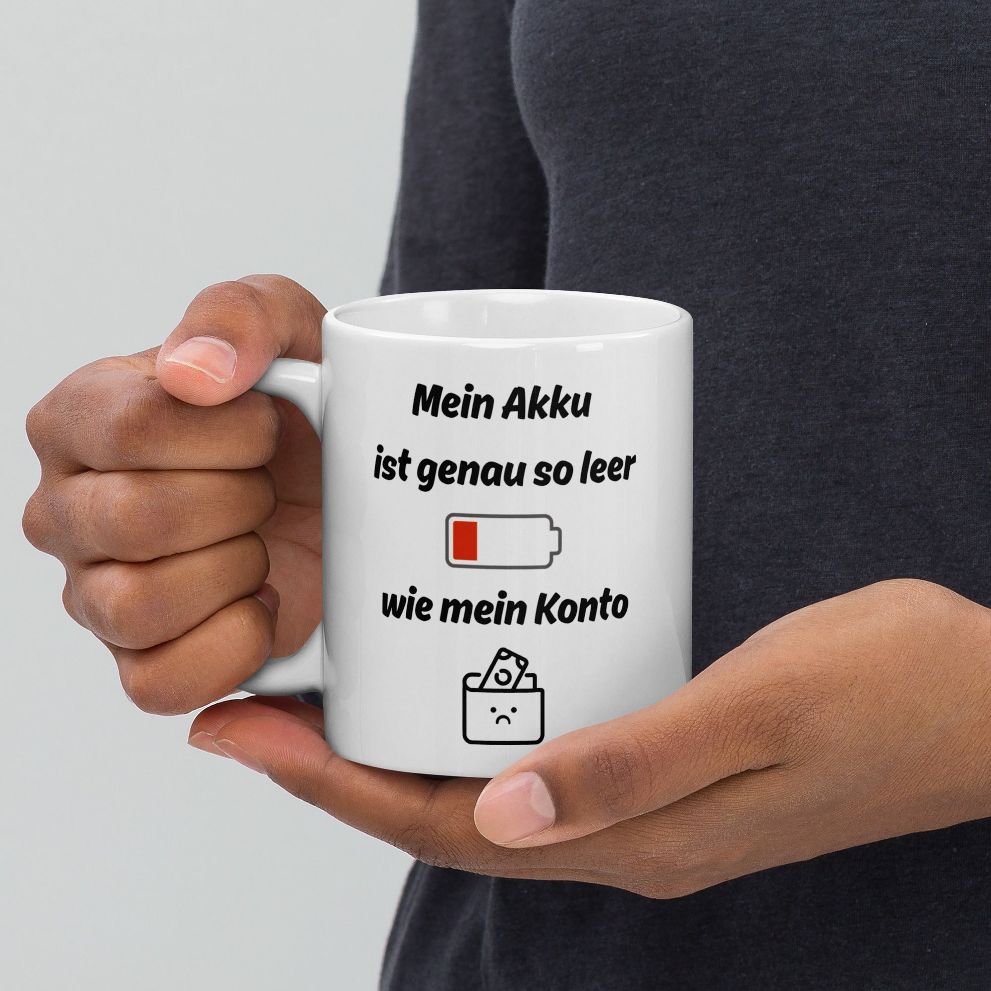Eine Hand hält eine weiße Kaffeetasse mit Spruch ‚Mein Akku ist genau so leer wie mein Konto‘ und Symbolen für leeren Akku und Geldbeutel.