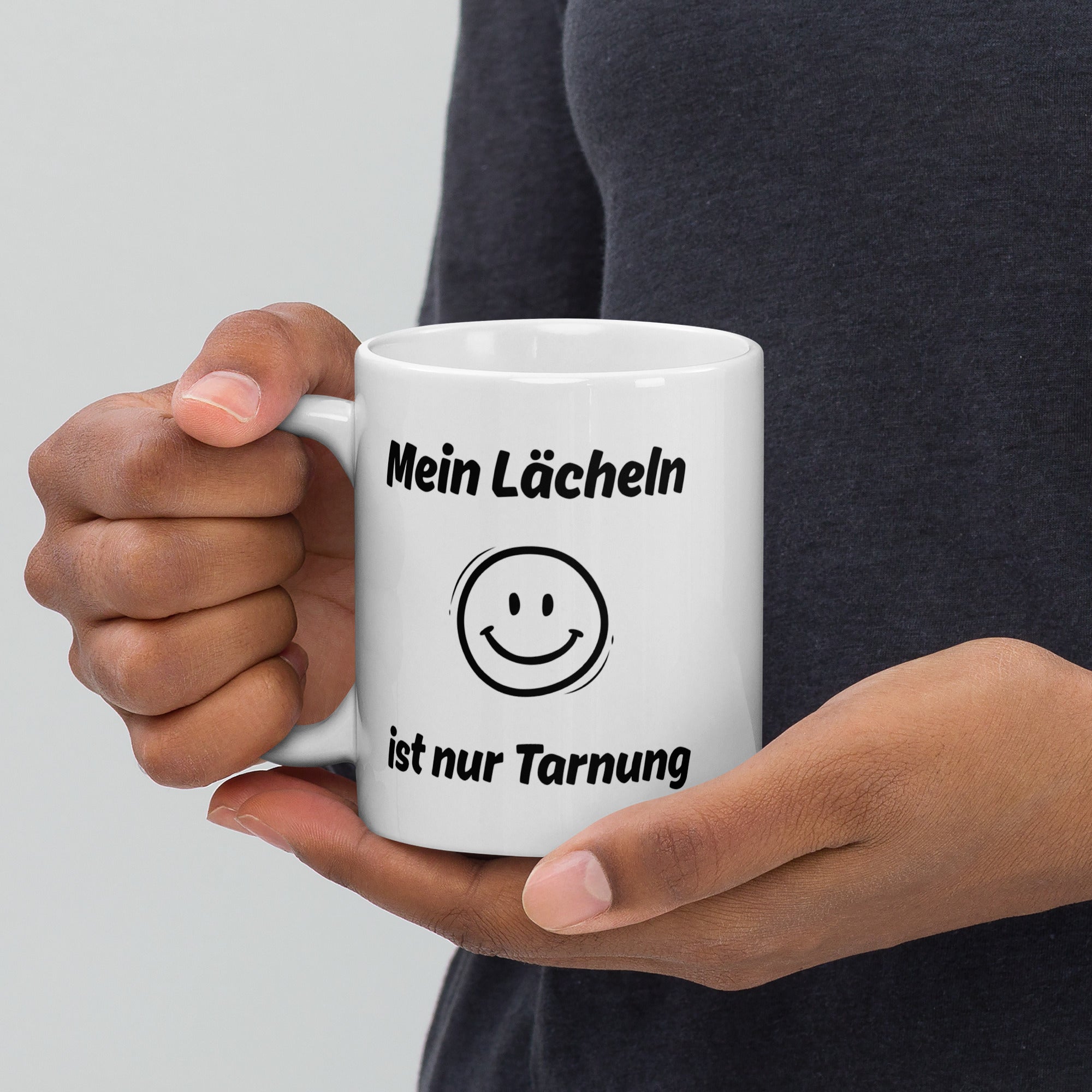 Eine Person hält eine weiße Kaffeetasse mit Spruch ‚Mein Lächeln ist nur Tarnung‘ und Smiley-Motiv.