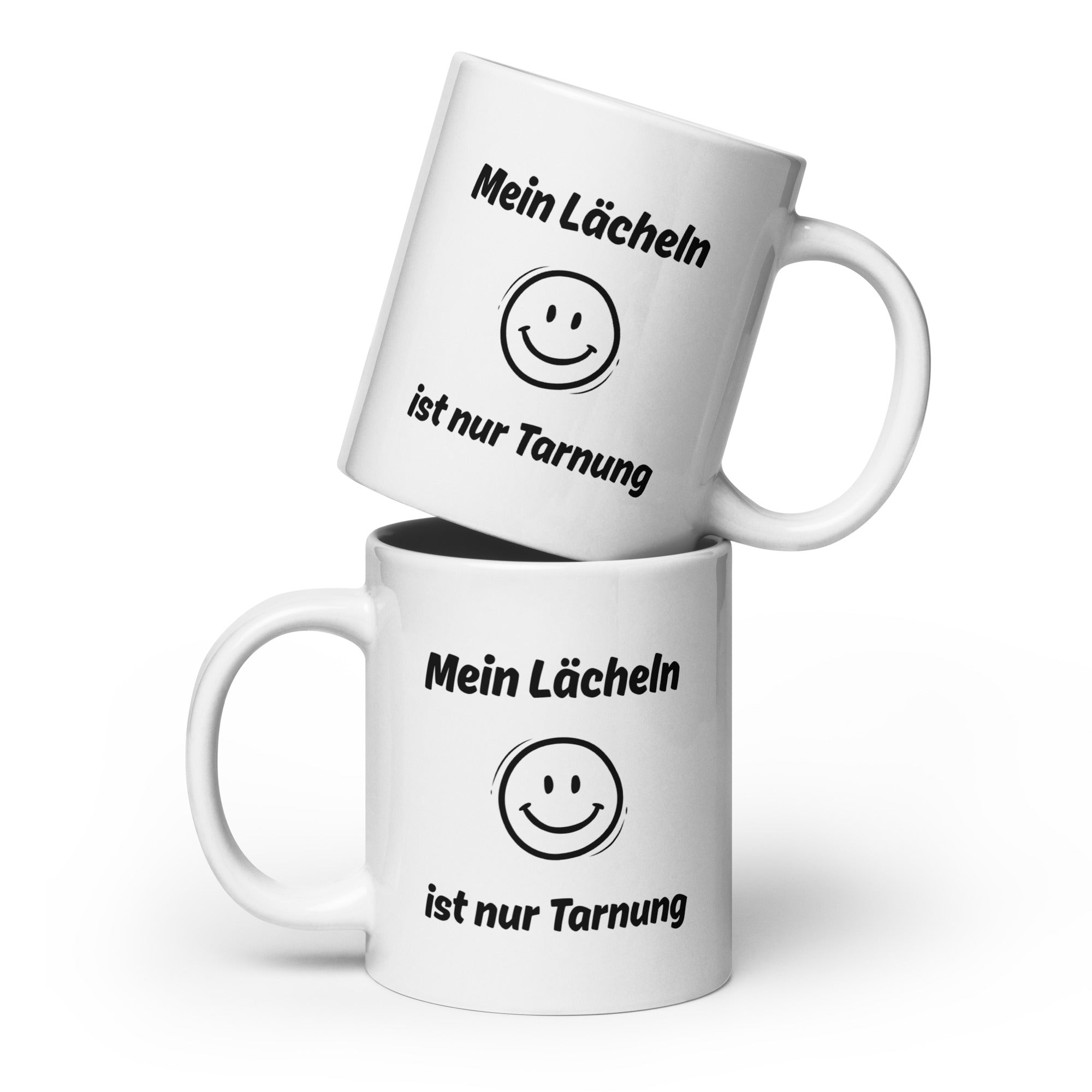 Zwei weiße Kaffeetassen mit schwarzer Aufschrift ‚Mein Lächeln ist nur Tarnung‘ und Smiley-Symbol, übereinandergestapelt.