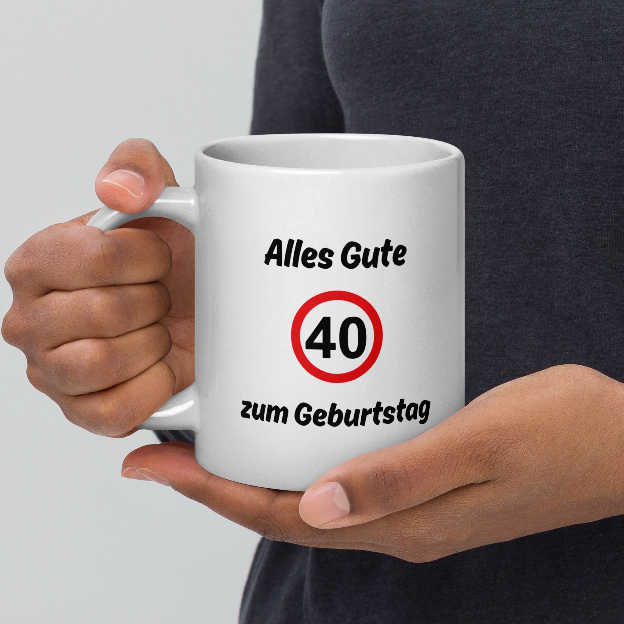 Eine Person hält eine weiße Kaffeetasse mit schwarzer Aufschrift ‚Alles Gute zum Geburtstag‘ und rotem Verkehrszeichen mit der Zahl 40.
